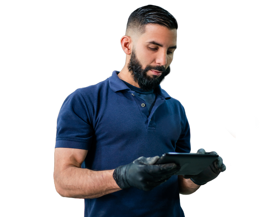 Mechanic in a navy polo uses a tablet while a car is on a lift in the background of a busy auto shop.