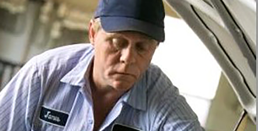 Male mechanic in a blue cap and striped shirt looking under the open hood of a car.