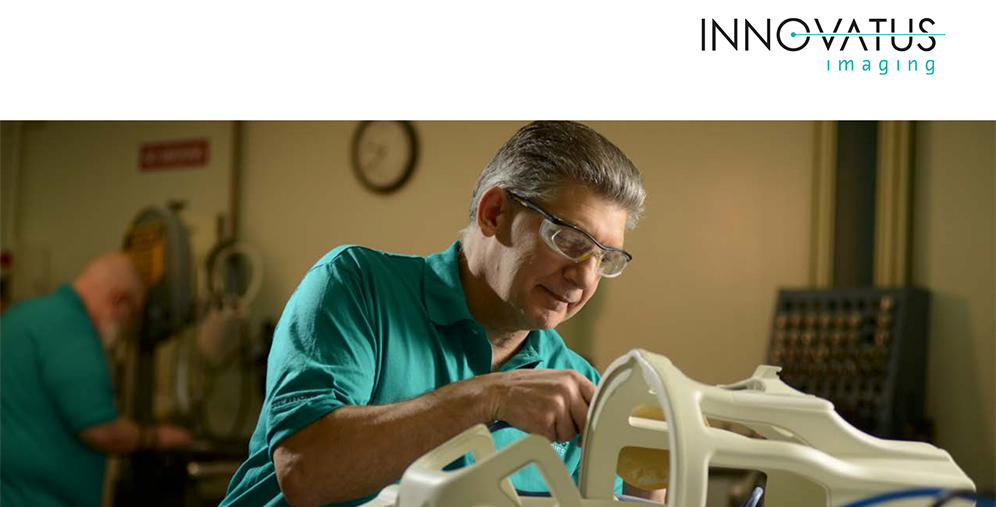 Technician wearing safety glasses inspects a white medical device frame on a workbench, with another worker in the background.